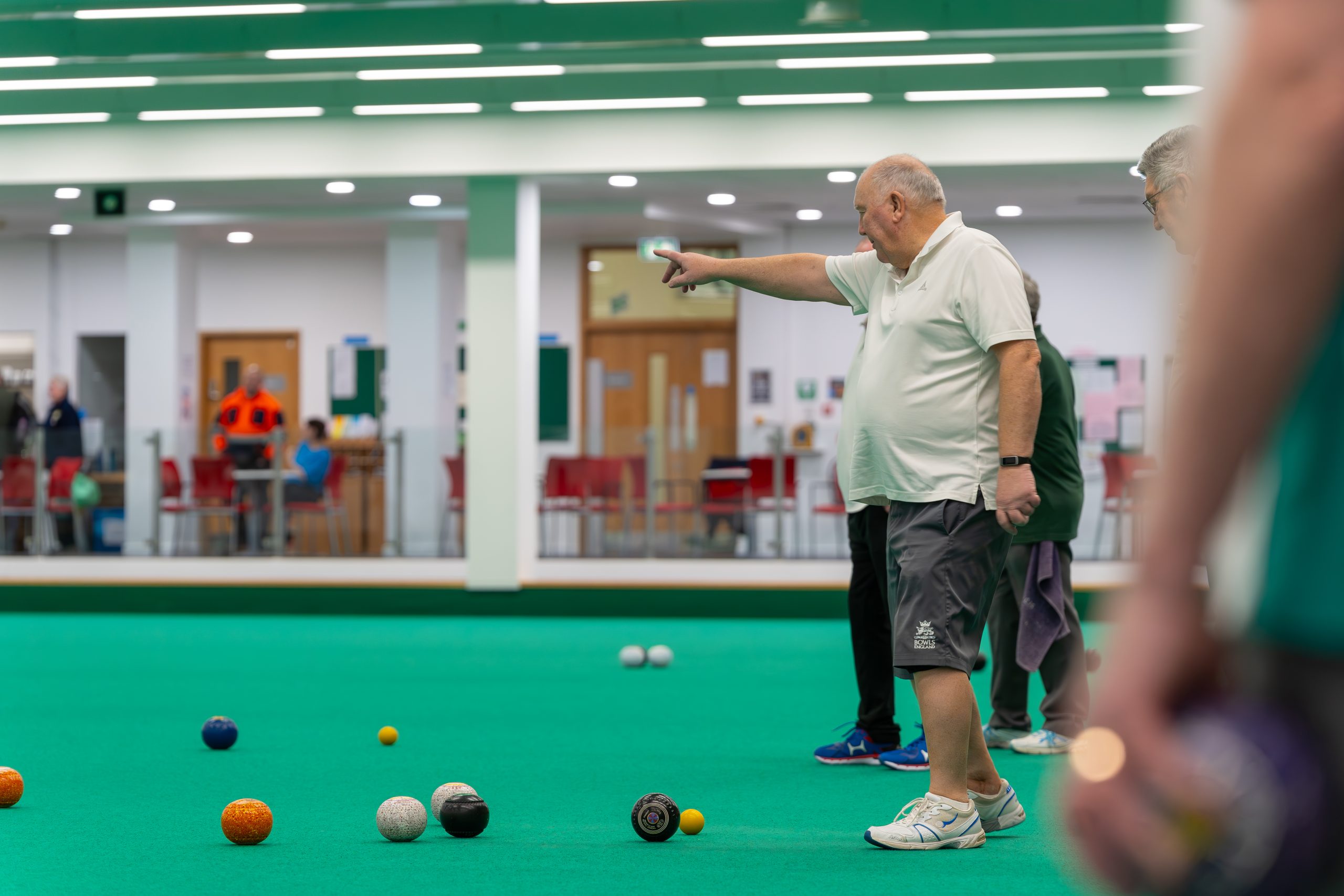 A man on the green bowls court pointing in the direction which his ball is rolling towards.