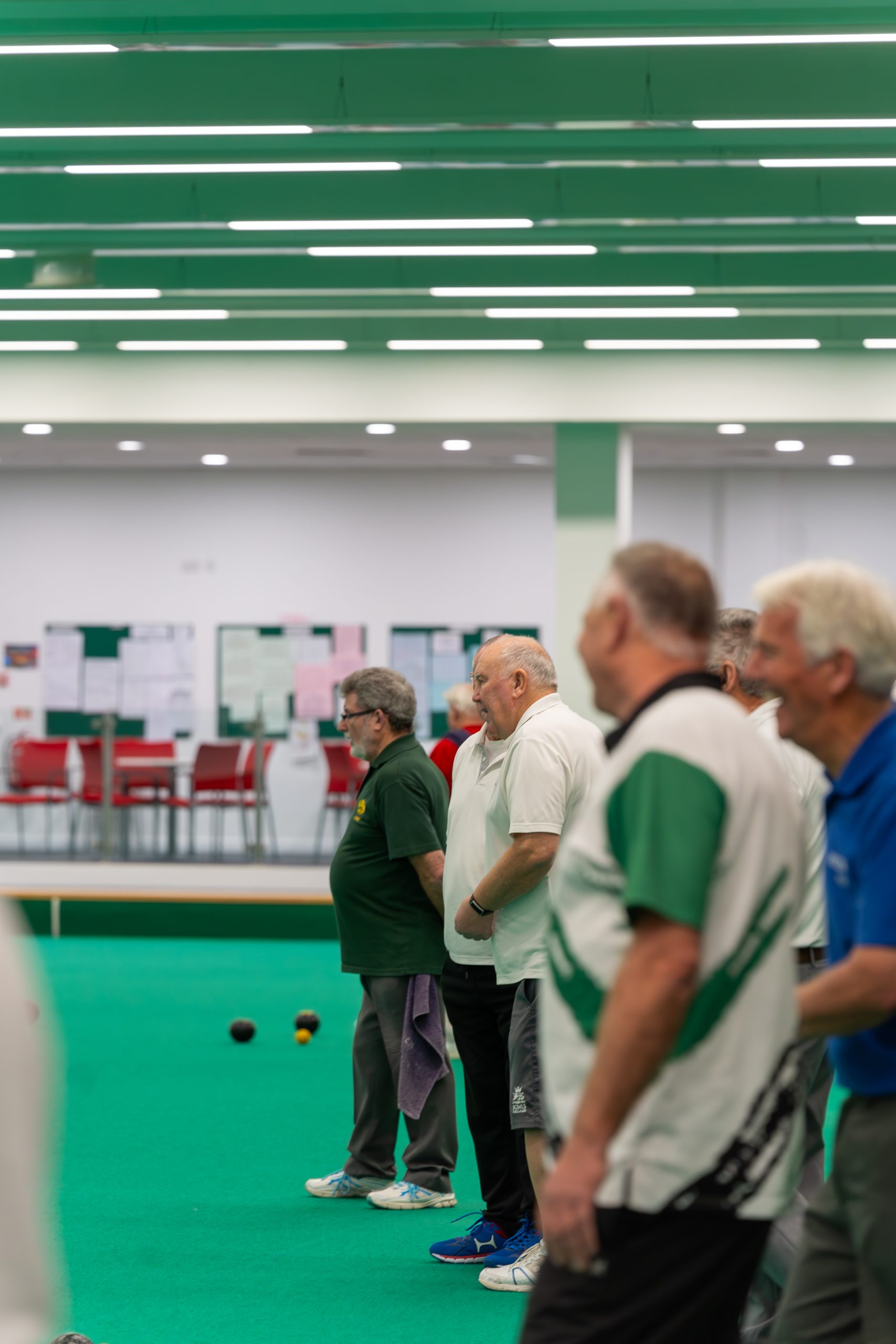 A group of men on a bowls rink watching another man bowl a ball down the green rink.